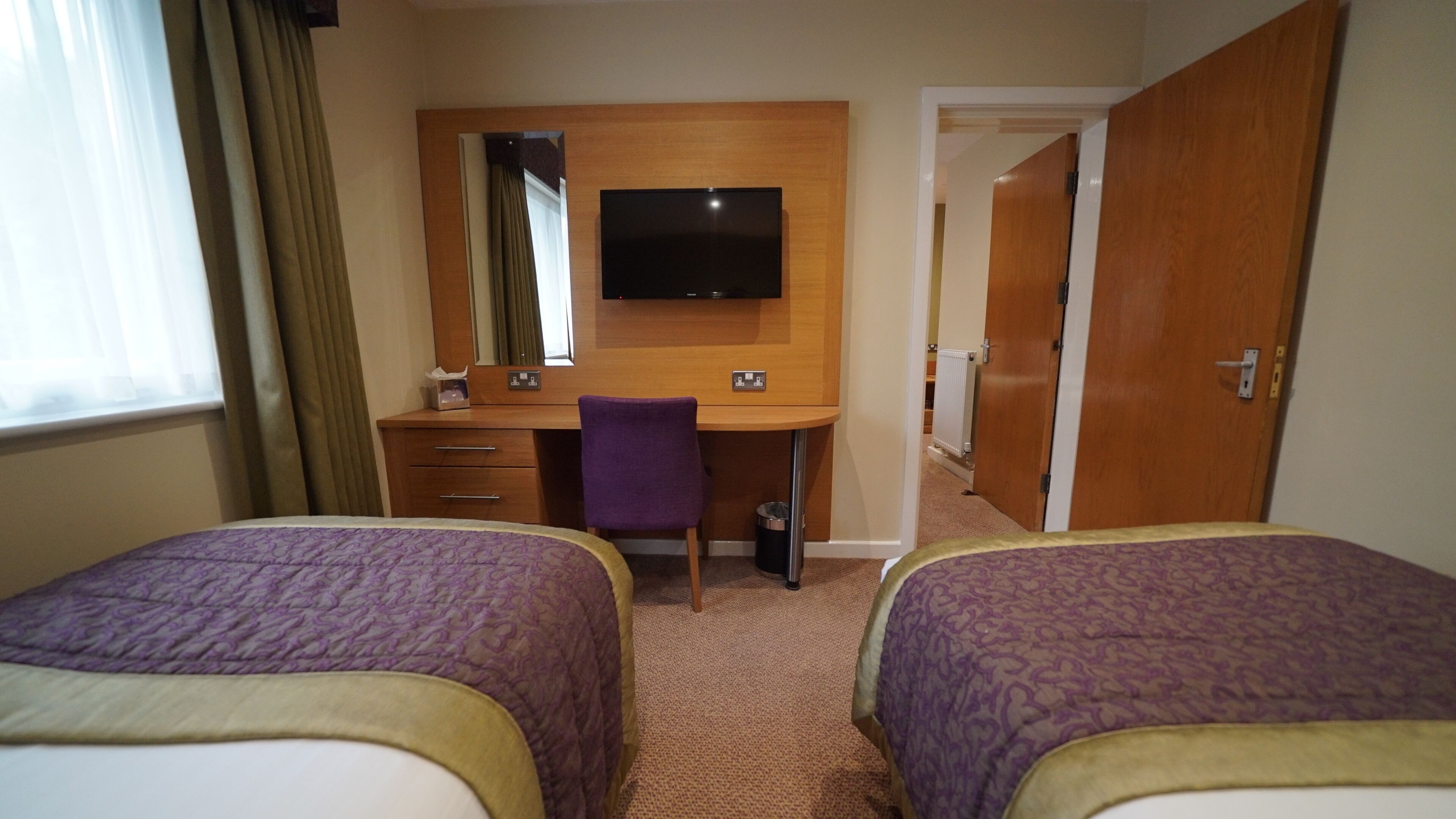 A hotel room with two beds, a desk with a chair, a television, and a door leading to another room.