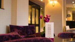 Uma área de recepção com cadeiras estofadas em roxo, uma placa branca listando tipos de eventos e uma luminária fixada na parede.