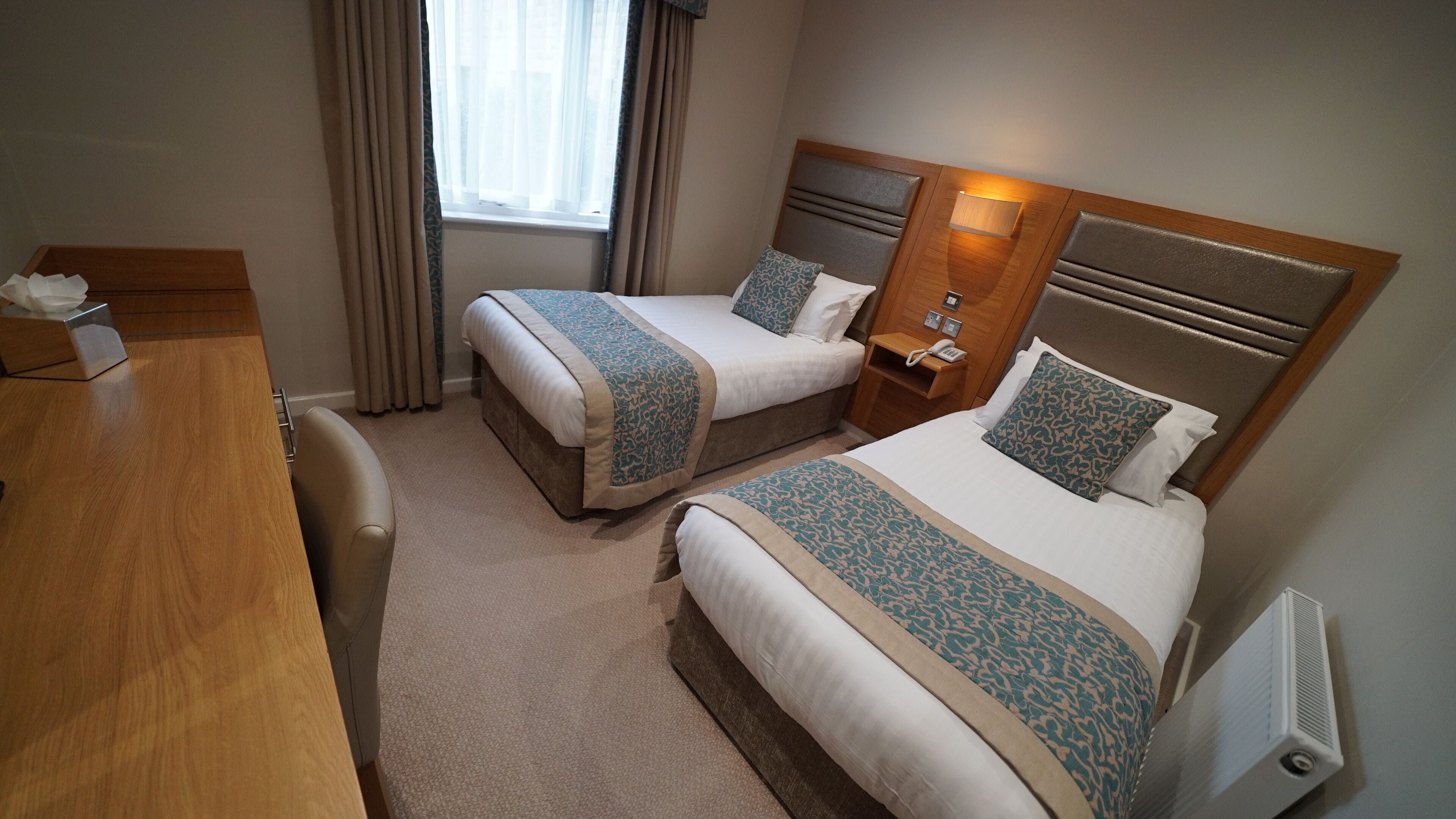 A hotel room with two beds, a wooden desk, a bedside table with a lamp, and a window with curtains.