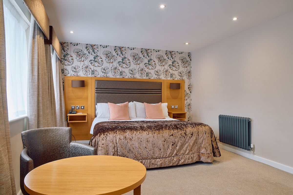 Una habitación de hotel con cama, una silla, una mesa, un radiador y una pared con papel tapiz floral.
