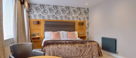 Una habitación de hotel con cama, una silla, una mesa, un radiador y una pared con papel tapiz floral.