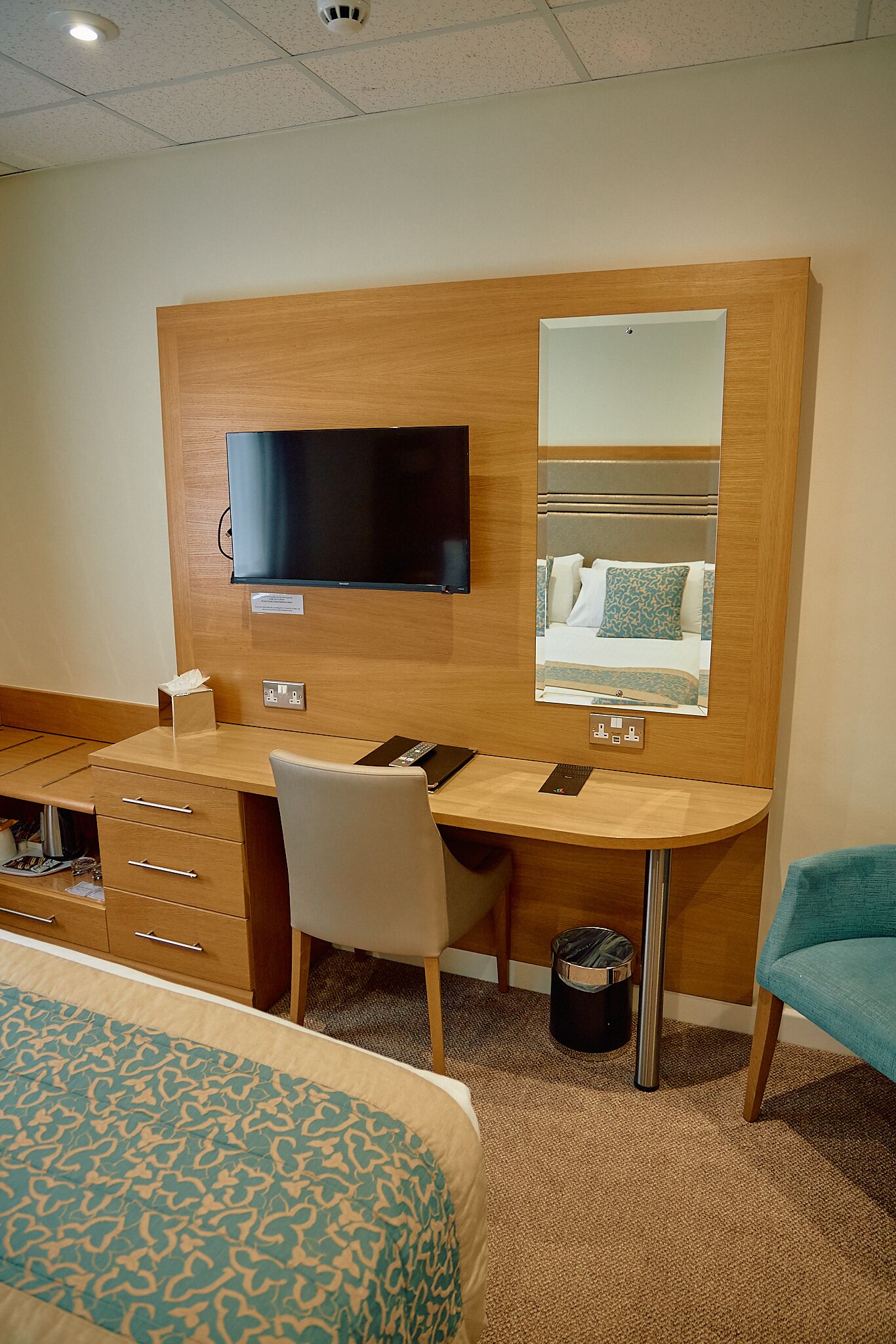 A hotel room with a wooden desk, a television, a mirror, a bed, and a trash can.