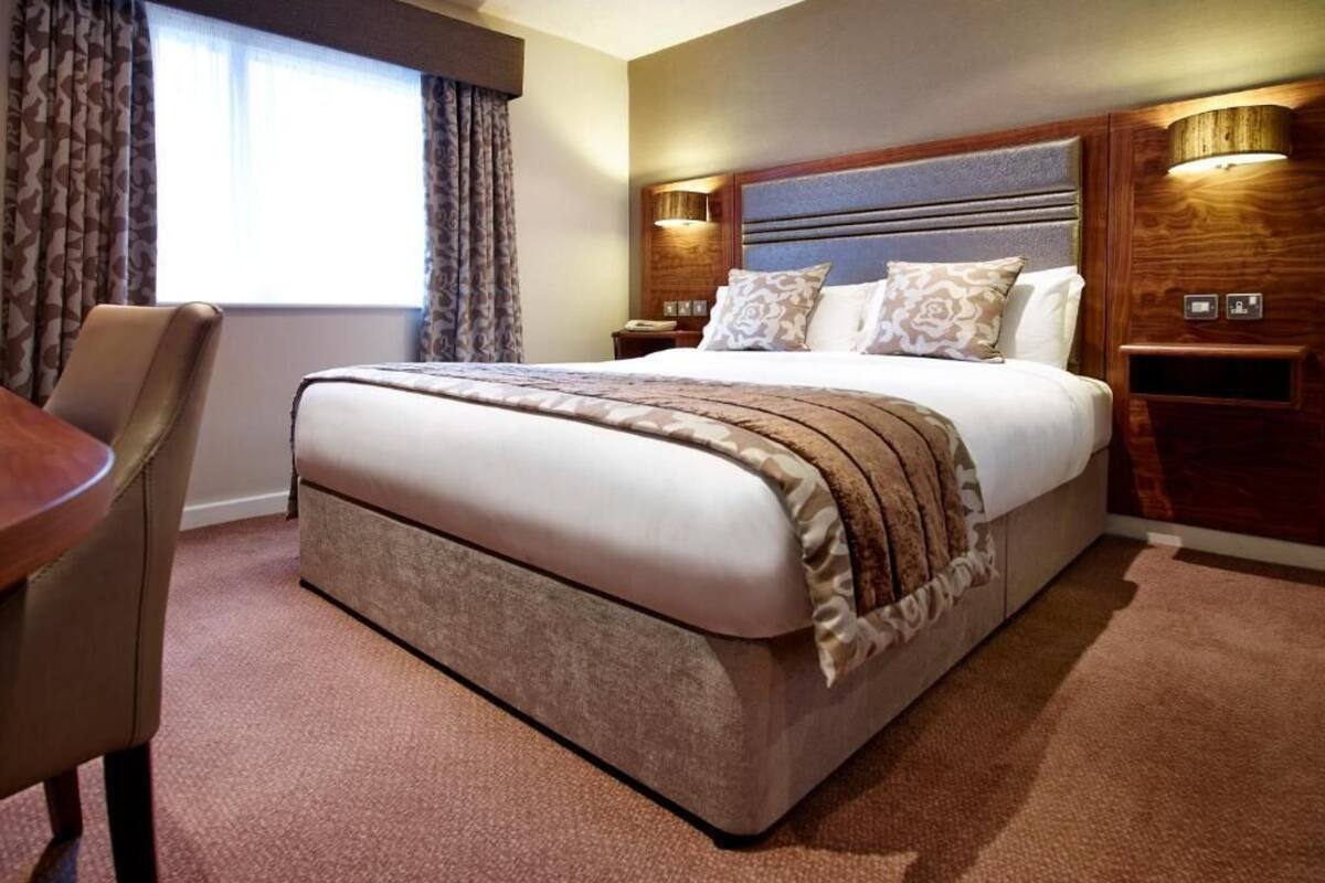 Una habitación de hotel con una cama grande, cabecera de madera, mesita de noche con lámpara y una silla.