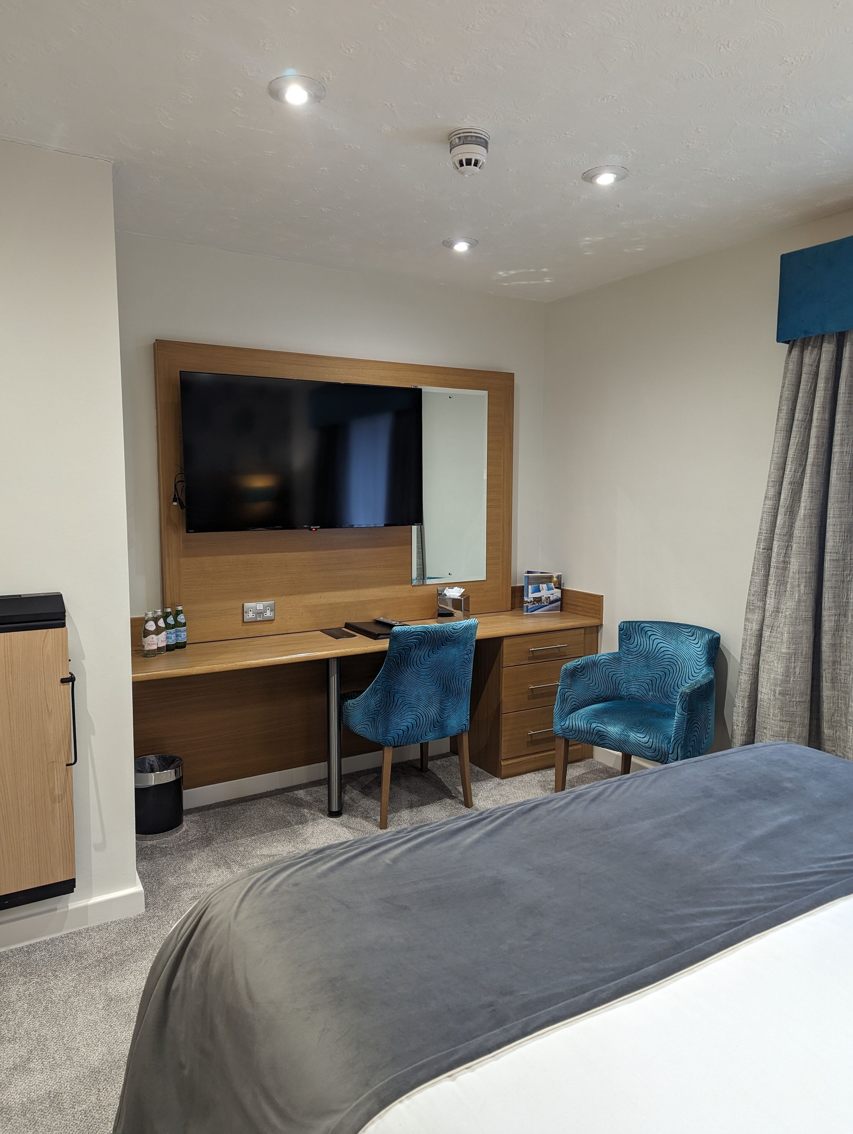 A hotel room with a bed, a desk with a flat-screen TV, two blue patterned chairs, a wooden dresser, and a trash can.