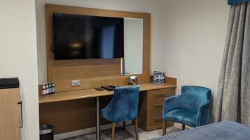 A hotel room with a bed, a desk with a flat-screen TV, two blue patterned chairs, a wooden dresser, and a trash can.