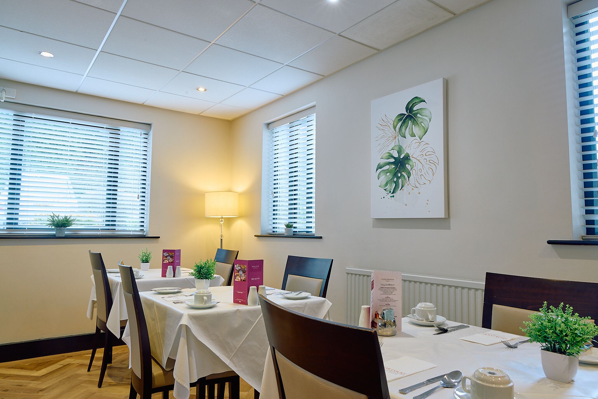 Comedor con mesa lista para cuatro, sillas, planta en maceta y lámpara de pared.