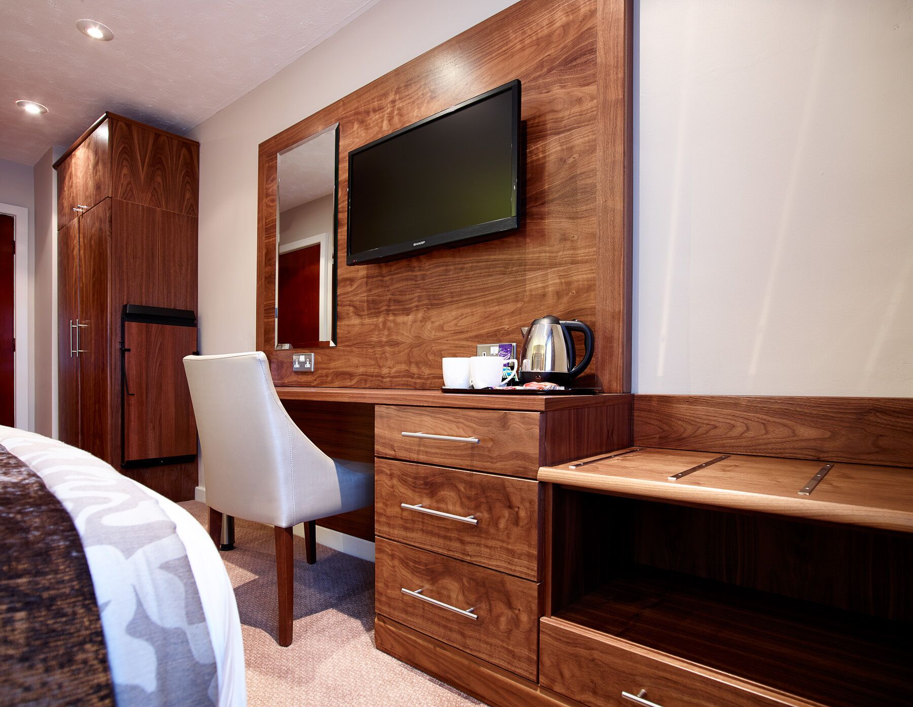 A hotel room with a wooden desk, a chair, a television mounted on the wall, and a minibar.