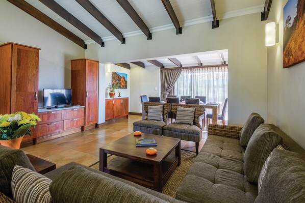 A spacious living room with a large sofa, a coffee table, and a television on a wooden cabinet. There is a dining area with a table and chairs in the background. - The Kingdom Resort (Moses Kotane)
