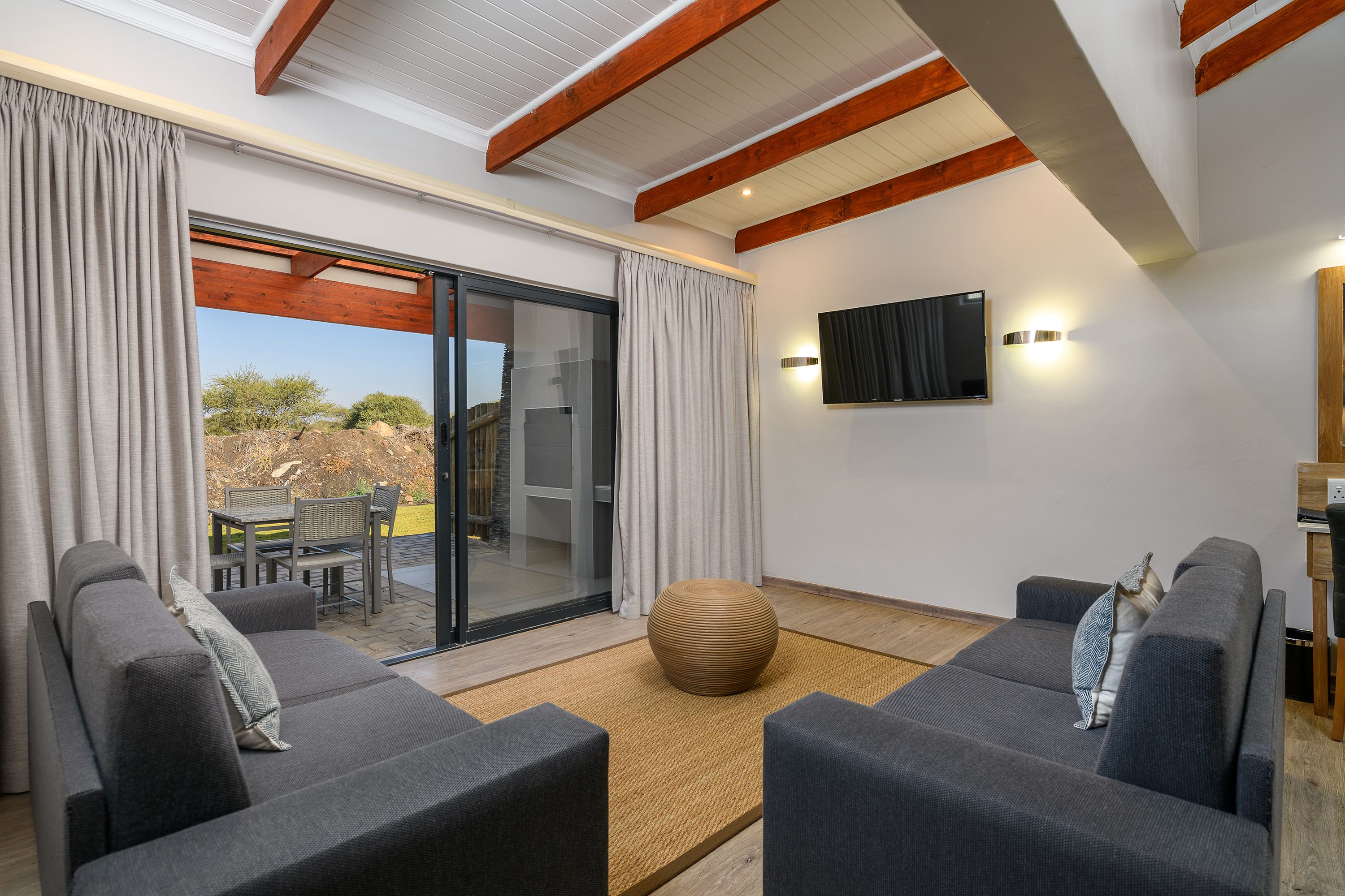 A modern living room with a sofa set, a flat-screen TV mounted on the wall, and a sliding glass door leading to an outdoor area.