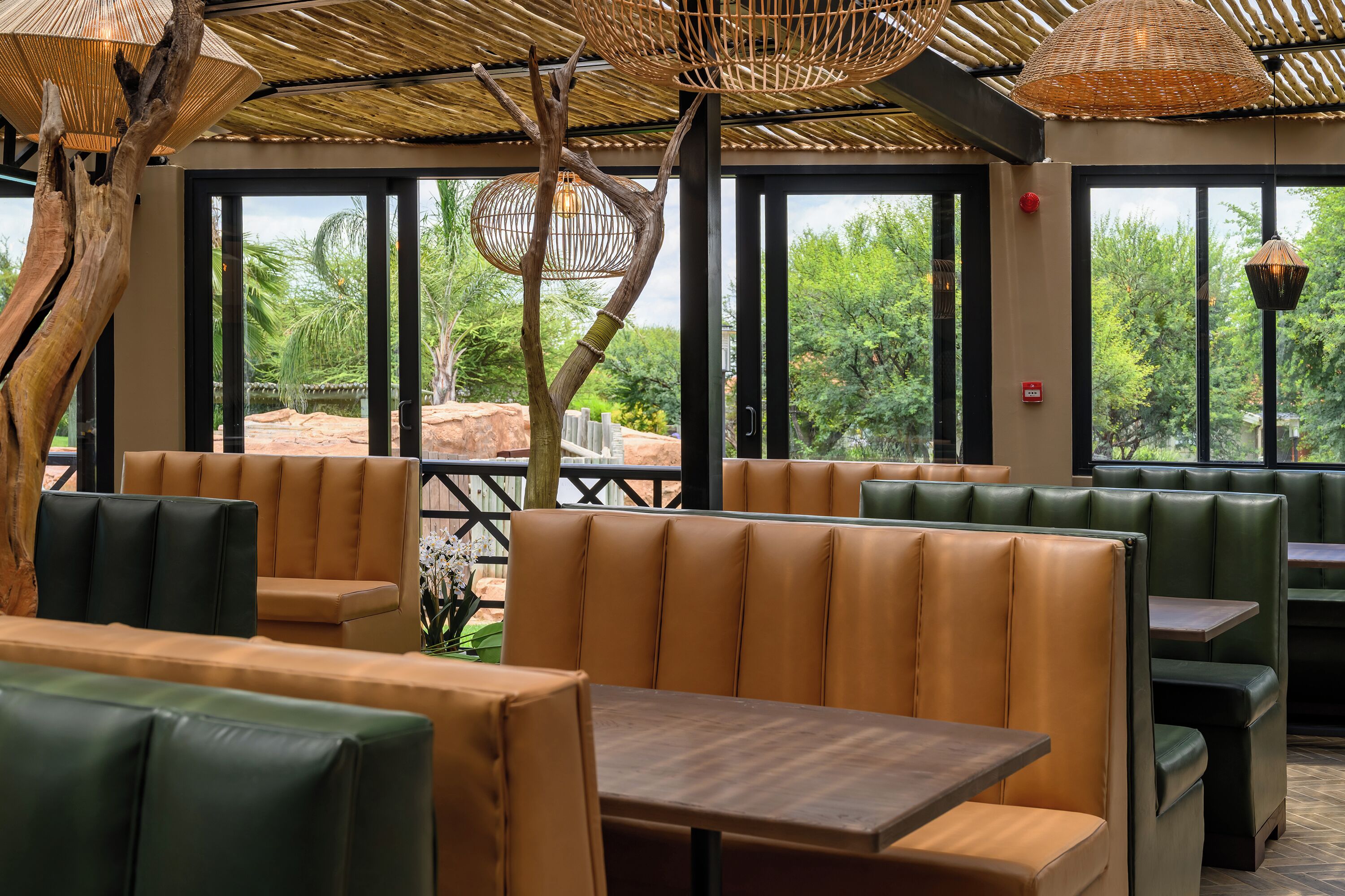 Ein Restaurant mit Ledersitzbänken und Tischen, großen Fenstern mit Blick auf Grünflächen und einer gewebten Decke.