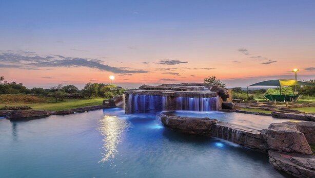 A waterfall cascading into a pool surrounded by rocks, with a sunset in the background. - The Kingdom Resort (Moses Kotane)