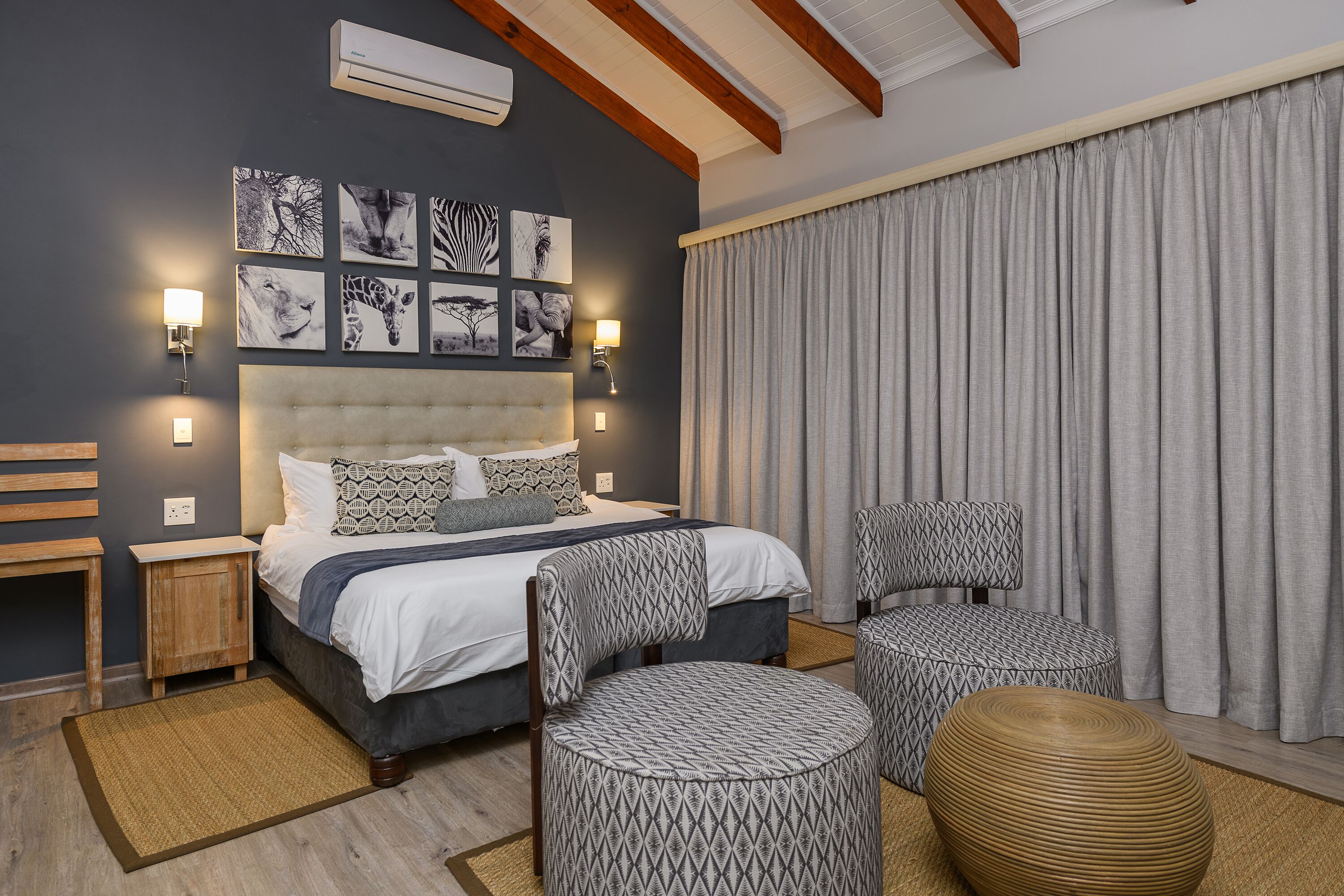 A bedroom with a large bed, two patterned chairs, a round ottoman, and wall-mounted artwork.
