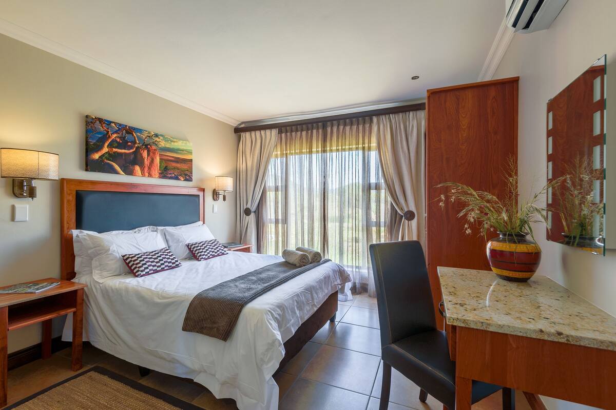 A hotel room with a large bed, a desk with a chair, a potted plant, and a painting on the wall.