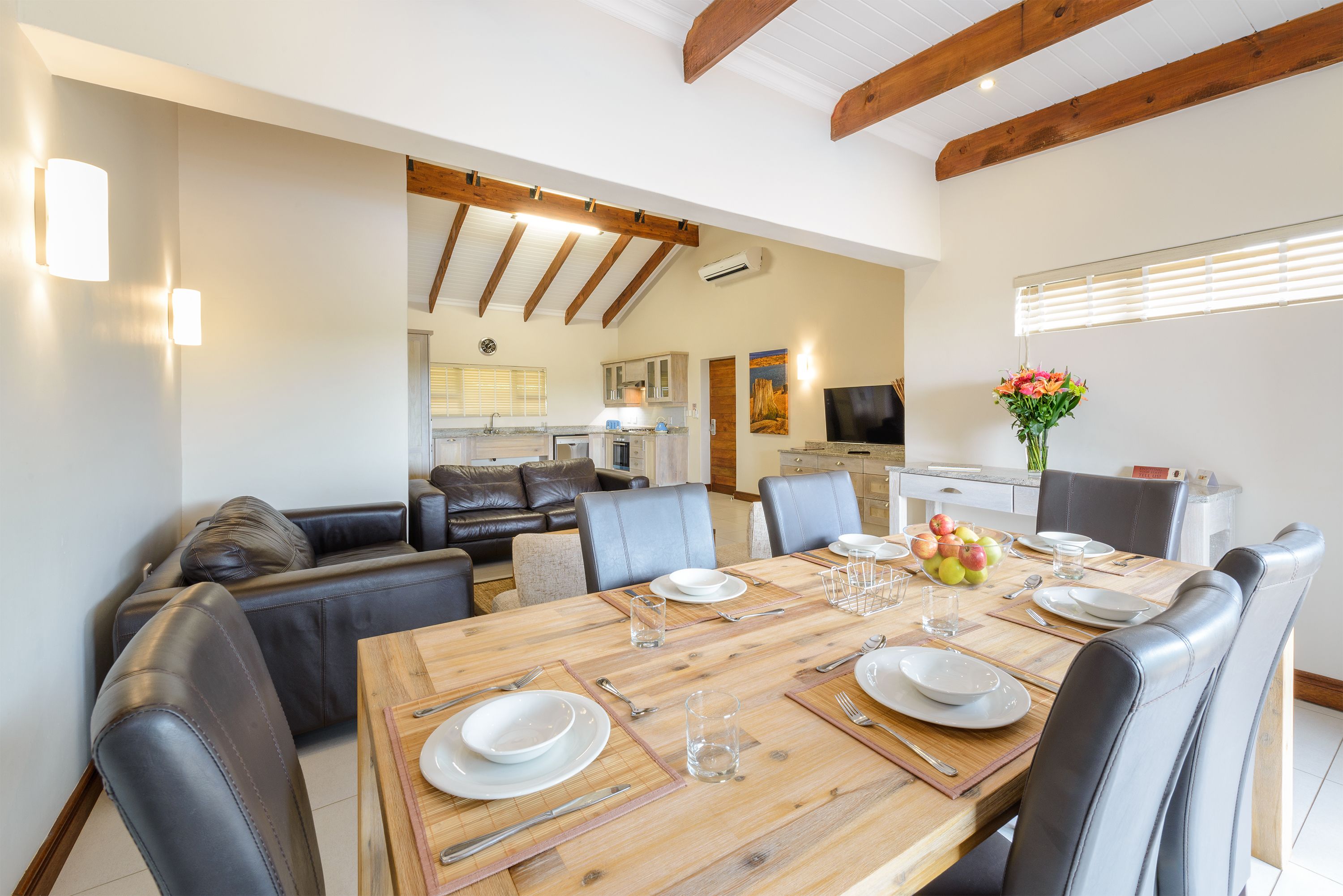 A dining area with a wooden table set for four, surrounded by leather chairs. A kitchen is visible in the background with modern appliances.