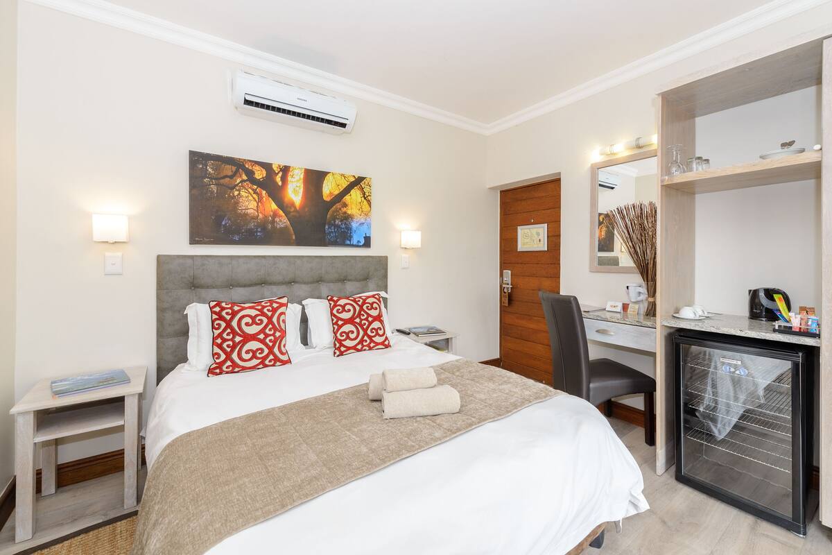 A hotel room with a bed, bedside tables, a desk, a refrigerator, and a mirror.