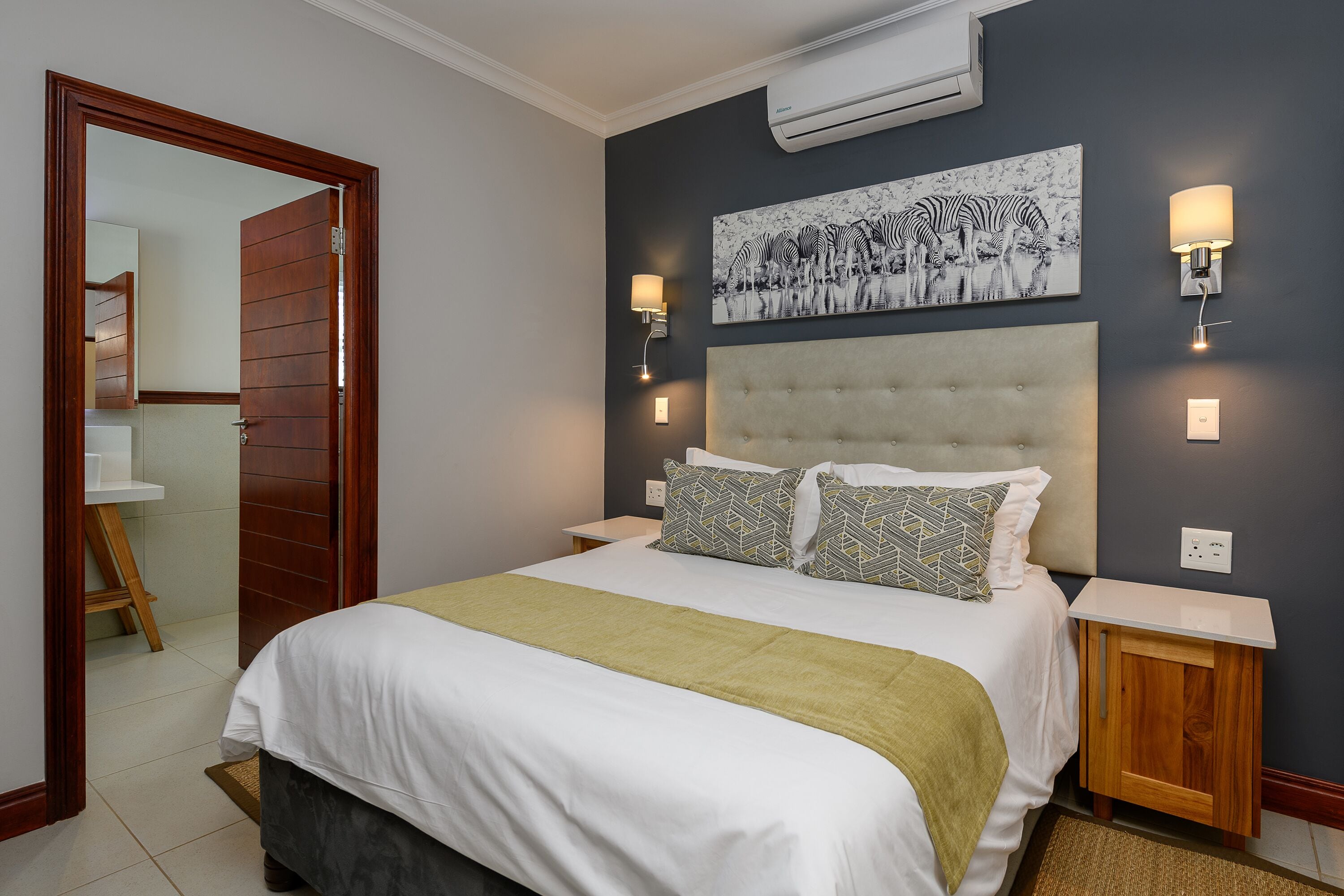 A bedroom with a large bed, a headboard, two bedside tables, a wall-mounted light fixture, and a wall-mounted air conditioner.