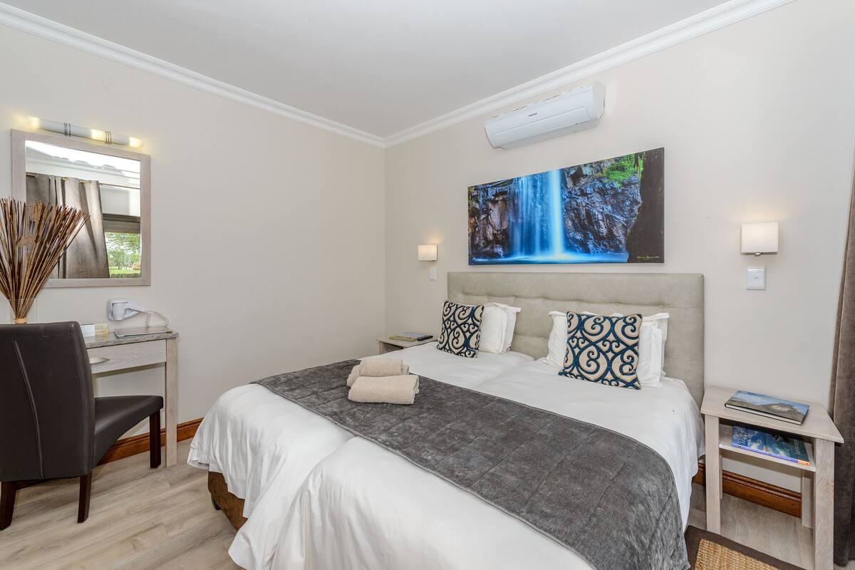 A hotel room with a large bed, a desk with a chair, a mirror, a painting of a waterfall, and a nightstand with a lamp.