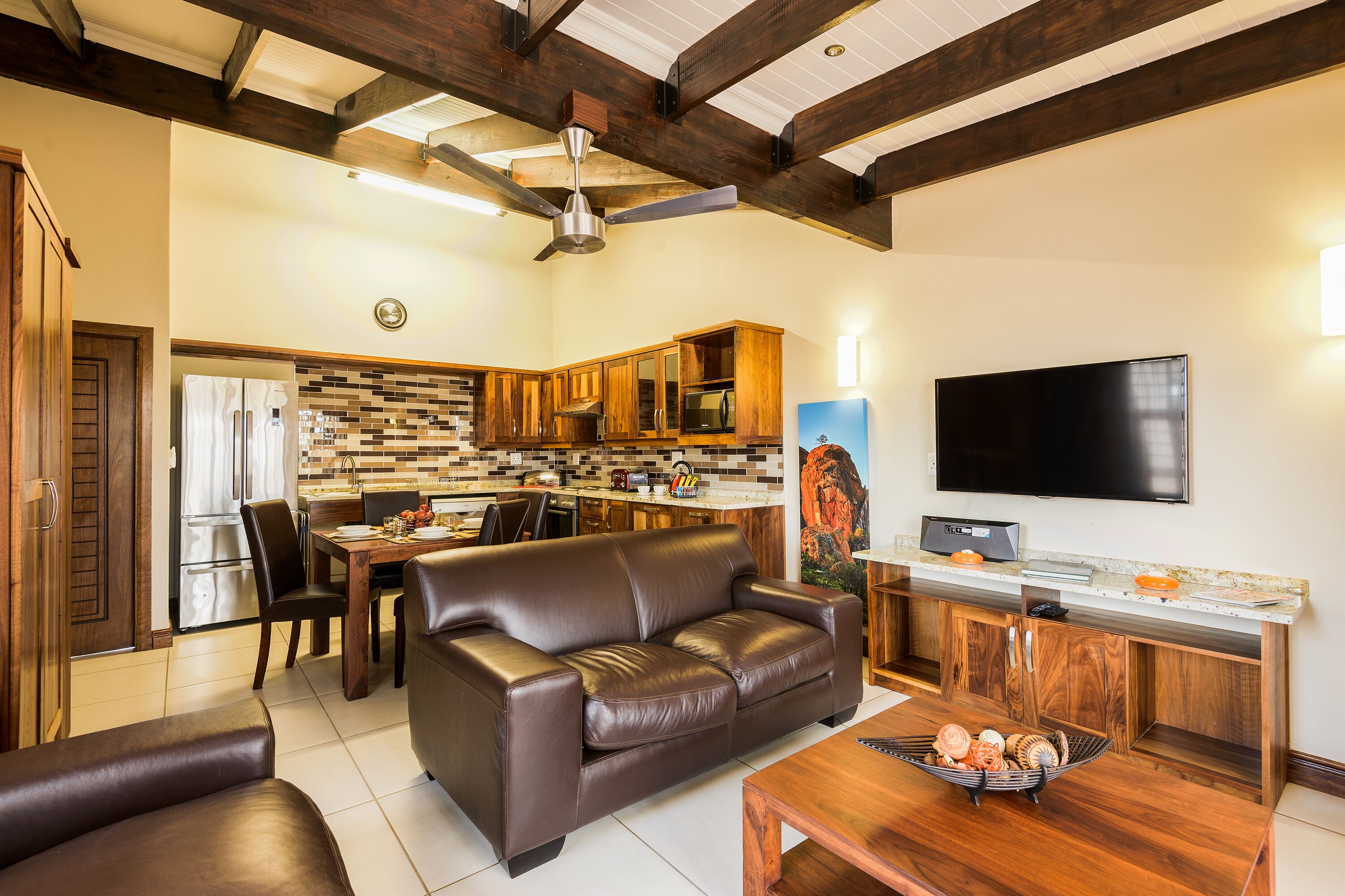 A modern living room with a leather sofa, a dining area with a wooden table, and a kitchen with wooden cabinets and a built-in microwave.