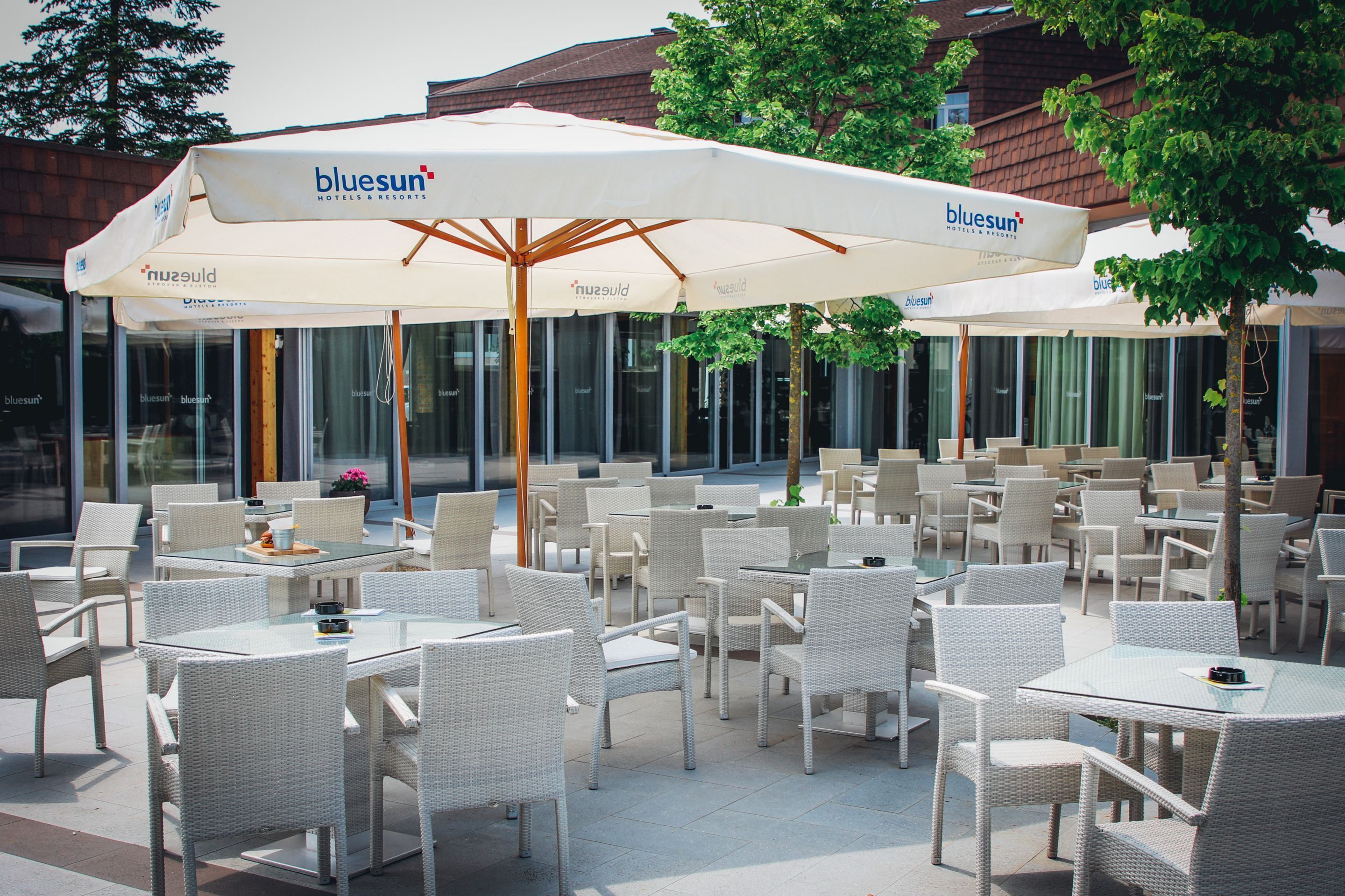 Un espace extérieur aménagé avec des chaises et des tables en osier blanc, protégé par un grand parasol arborant le logo « bluesun ».