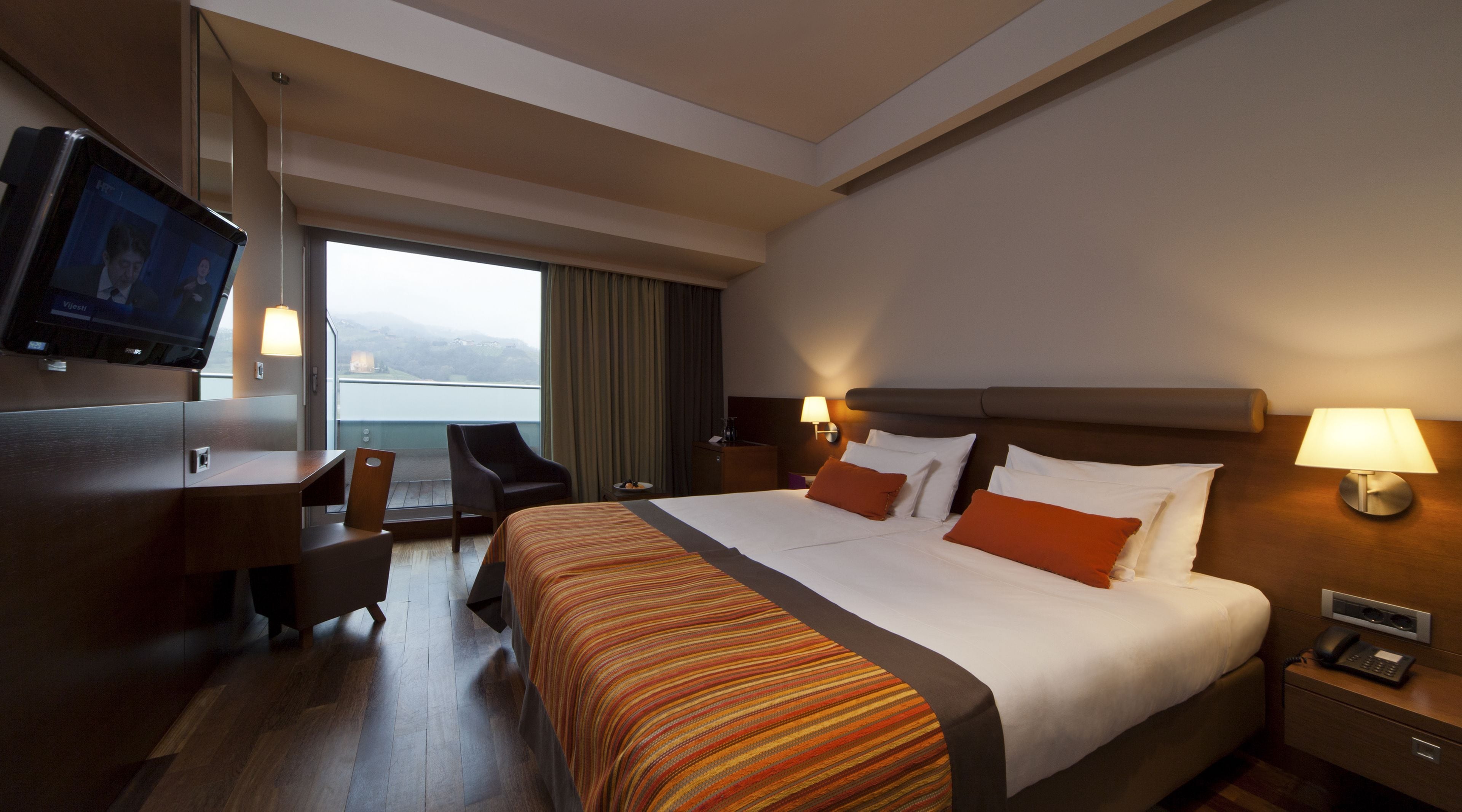 Ein Hotelzimmer mit einem großen Bett, einem Schreibtisch, einem Sessel, einem Fernseher und Blick ins Freie.