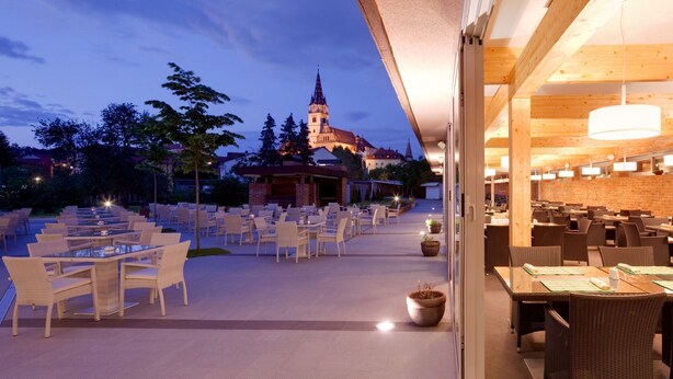An outdoor dining area with tables and chairs, a brick wall, and a view of a church tower. - Hotel Kaj (Marija Bistrica)