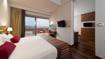 A hotel room with a large bed, a desk, a chair, a TV, and a balcony with a view.