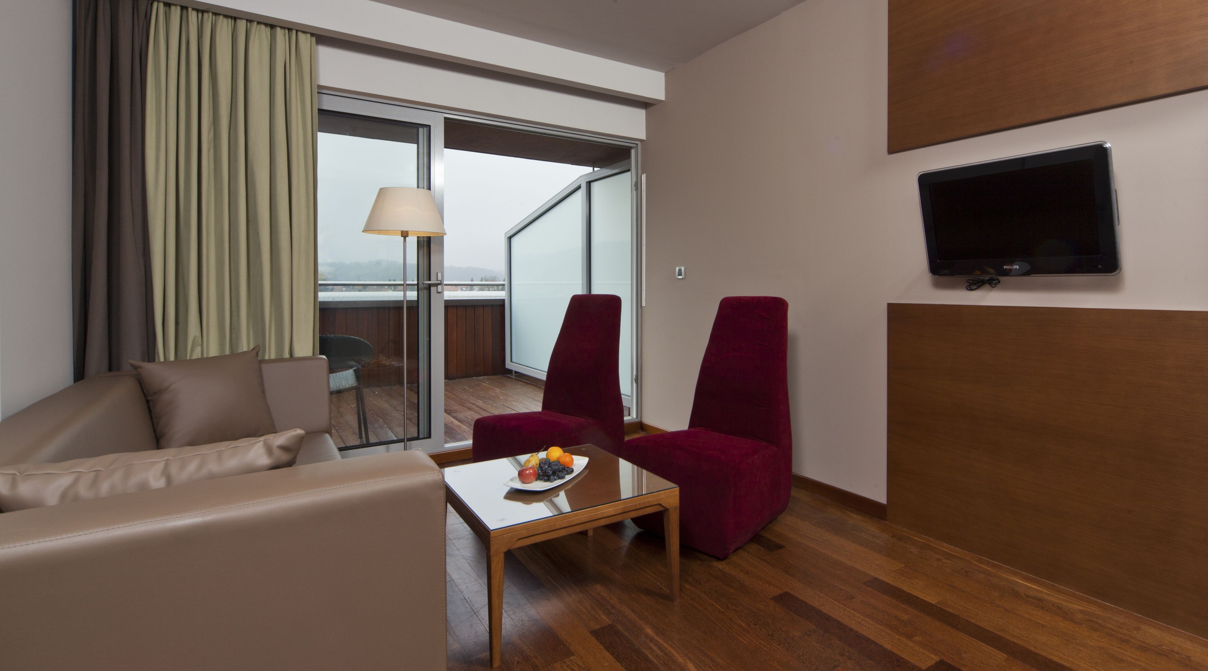 Ein Hotelzimmer mit einer Couch, zwei roten Sesseln, einem kleinen Tisch mit Früchten, einem Fernseher und einem Balkon mit Blick.