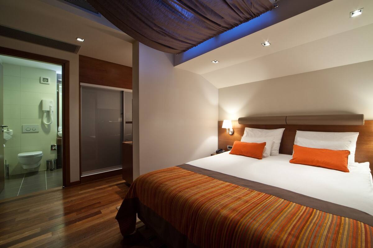 A modern hotel room with a large bed, two bedside lamps, a nightstand, and a bathroom visible through a glass partition.