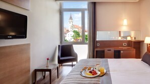 A hotel room with a bed, bedside table, chair, TV, and a view of a building through the window. - Hotel Kaj (Marija Bistrica)