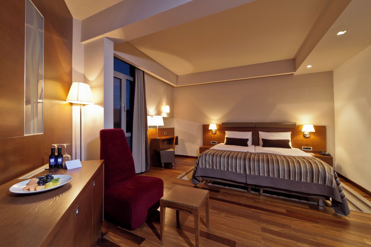 A hotel room with a bed, a desk with a plate of fruit, a red chair, and a small table.