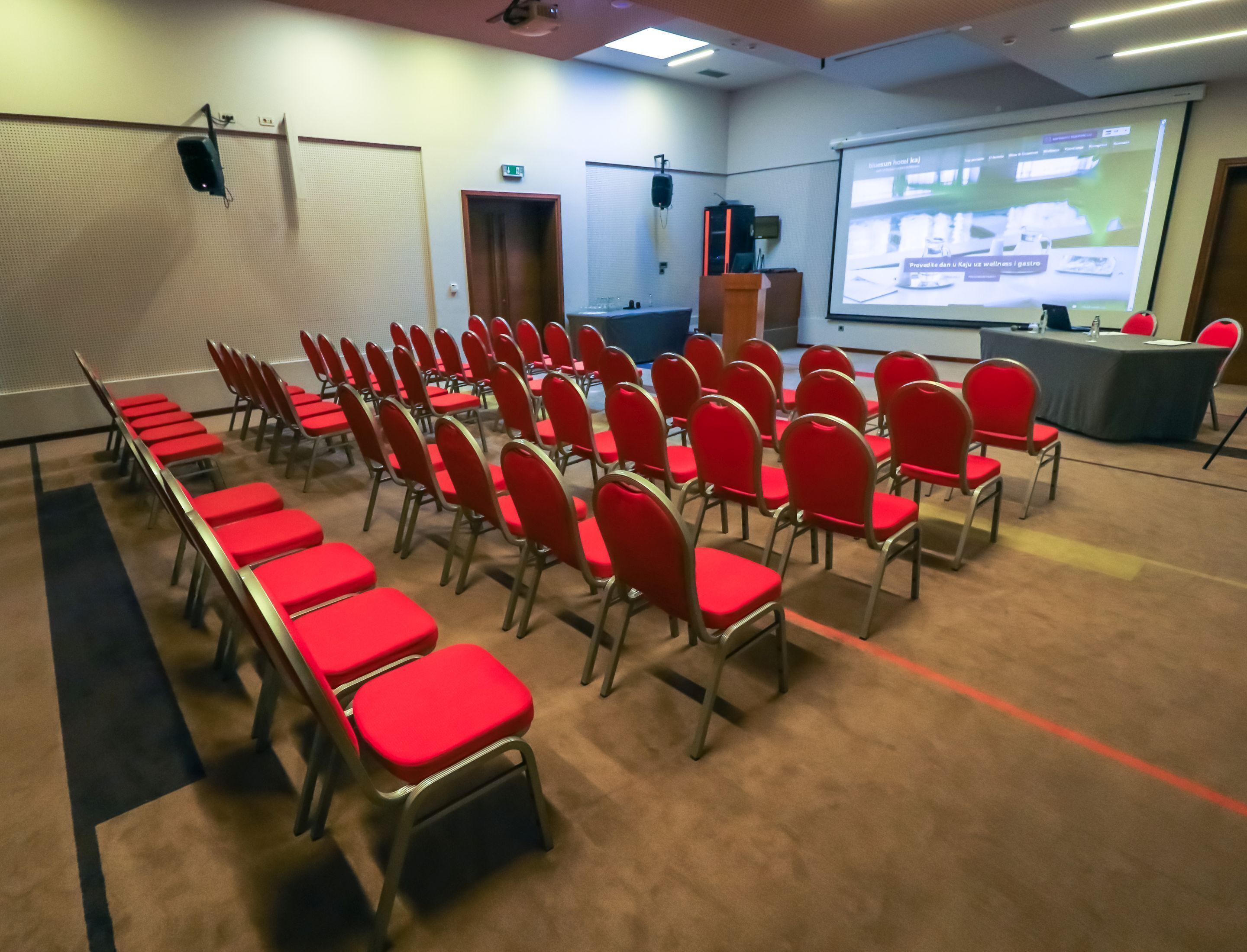 A conference room with rows of red chairs, a podium, and a projection screen displaying hotel information.