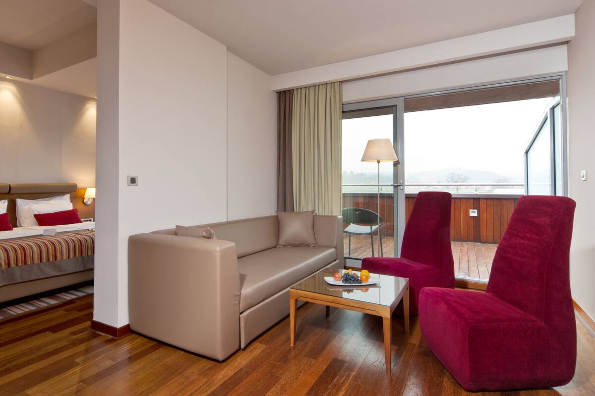 A hotel room with a bed, a sofa, a red armchair, a glass coffee table, and a view of the outdoors.
