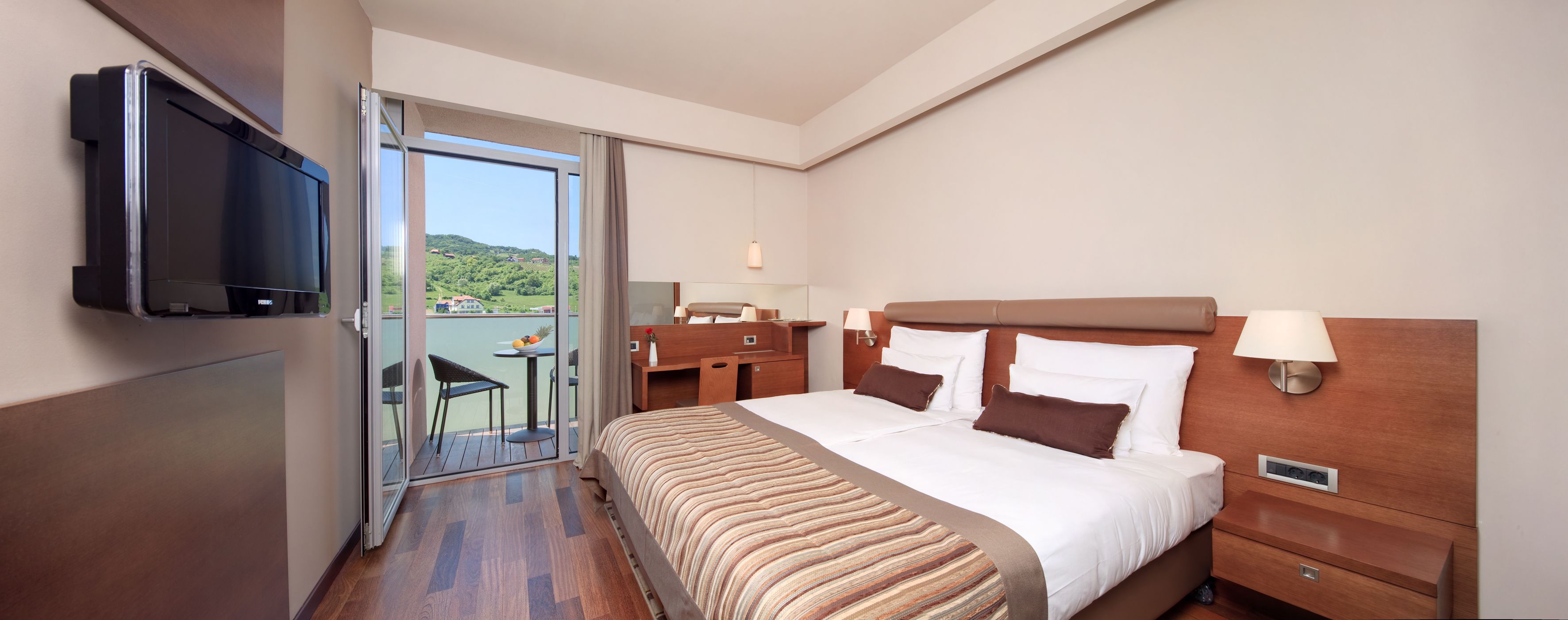 A hotel room with a bed, bedside tables, a television, a balcony with a table and chairs, and a view of greenery.