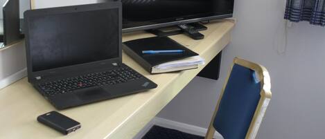 A hotel room with a desk, a laptop, a notebook, a smartphone, and a television.
