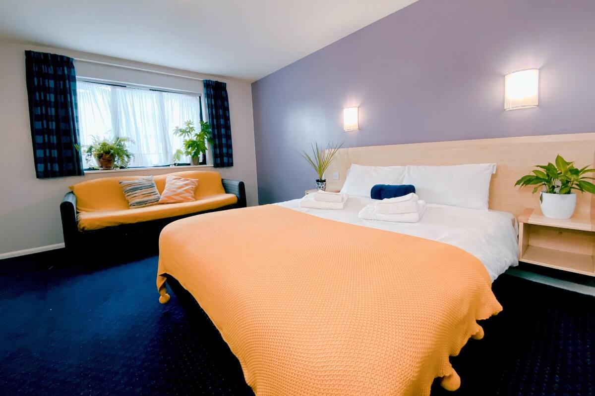 Ein Hotelzimmer mit einem großen Bett, einer Couch, zwei Zimmerpflanzen und einem Vorhangfenster.