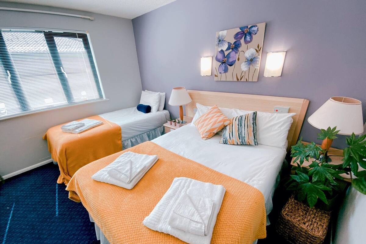 Ein Hotelzimmer mit zwei Betten, jedes mit einer weißen und orangefarbenen Decke, einem Kopfteil, Nachttischlampen, einem Wandbild mit Blumen und einer Topfpflanze.