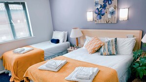 Una habitación de hotel con dos camas, cada una con edredón blanco y manta naranja. Hay dos mesitas de noche con lámparas, una luz en la pared y un cuadro de flores.