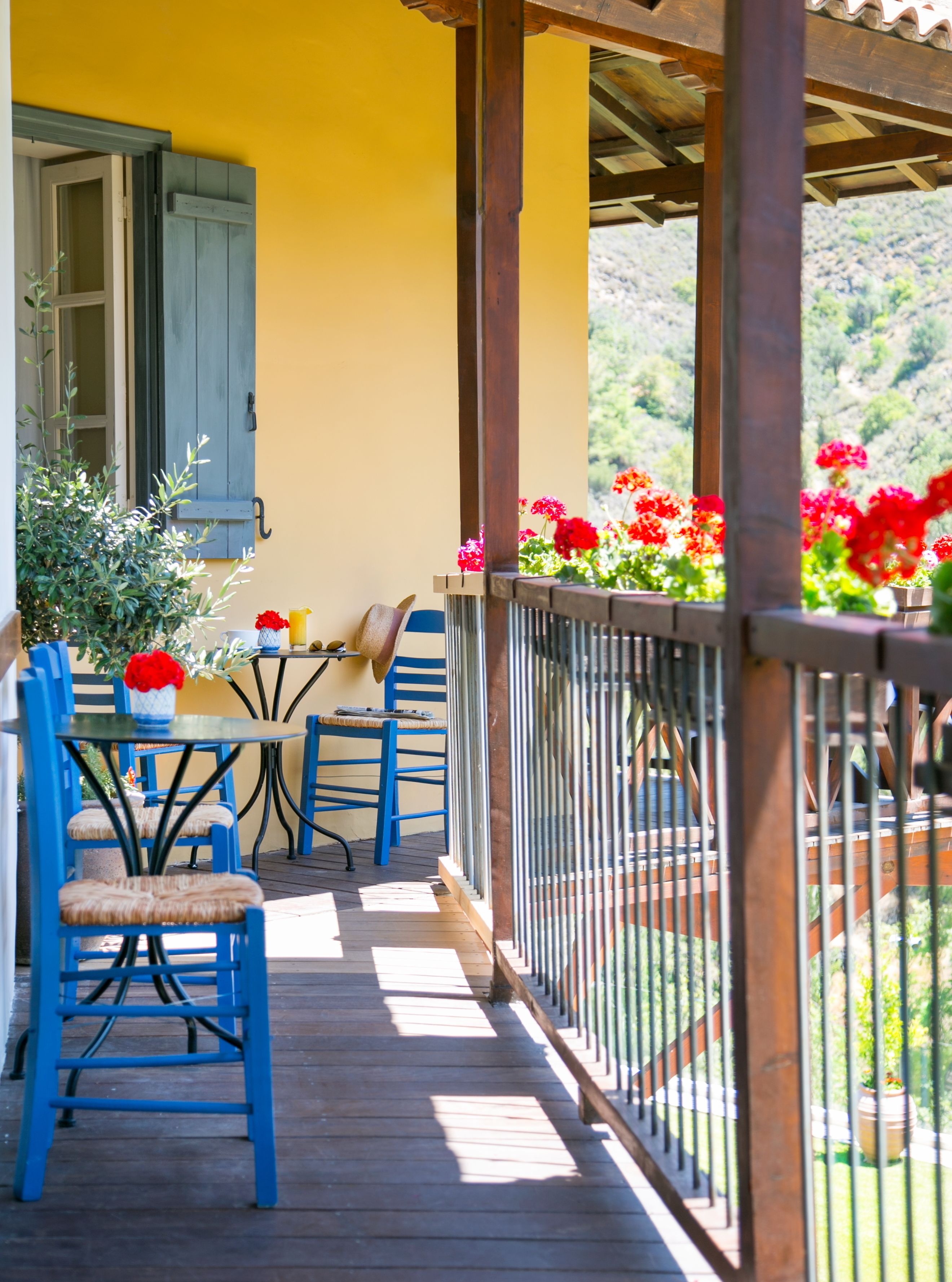 Eine Holzterrasse mit blauen Stühlen und einem Tisch, eine gelbe Wand und ein Geländer mit roten Blumen.