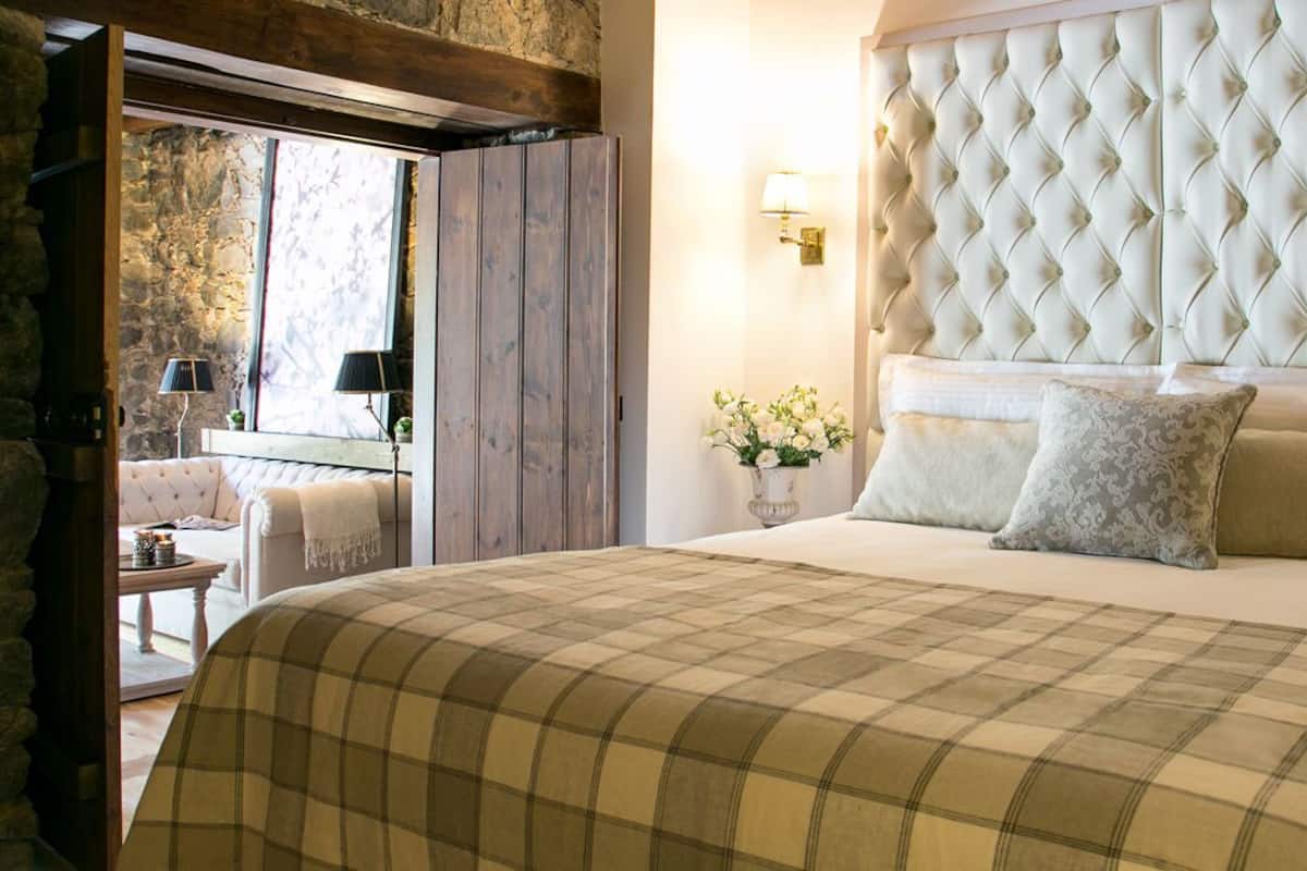 A bedroom with a large bed, a tufted headboard, a checkered bedspread, a nightstand with a lamp, and a view of a stone wall and balcony.