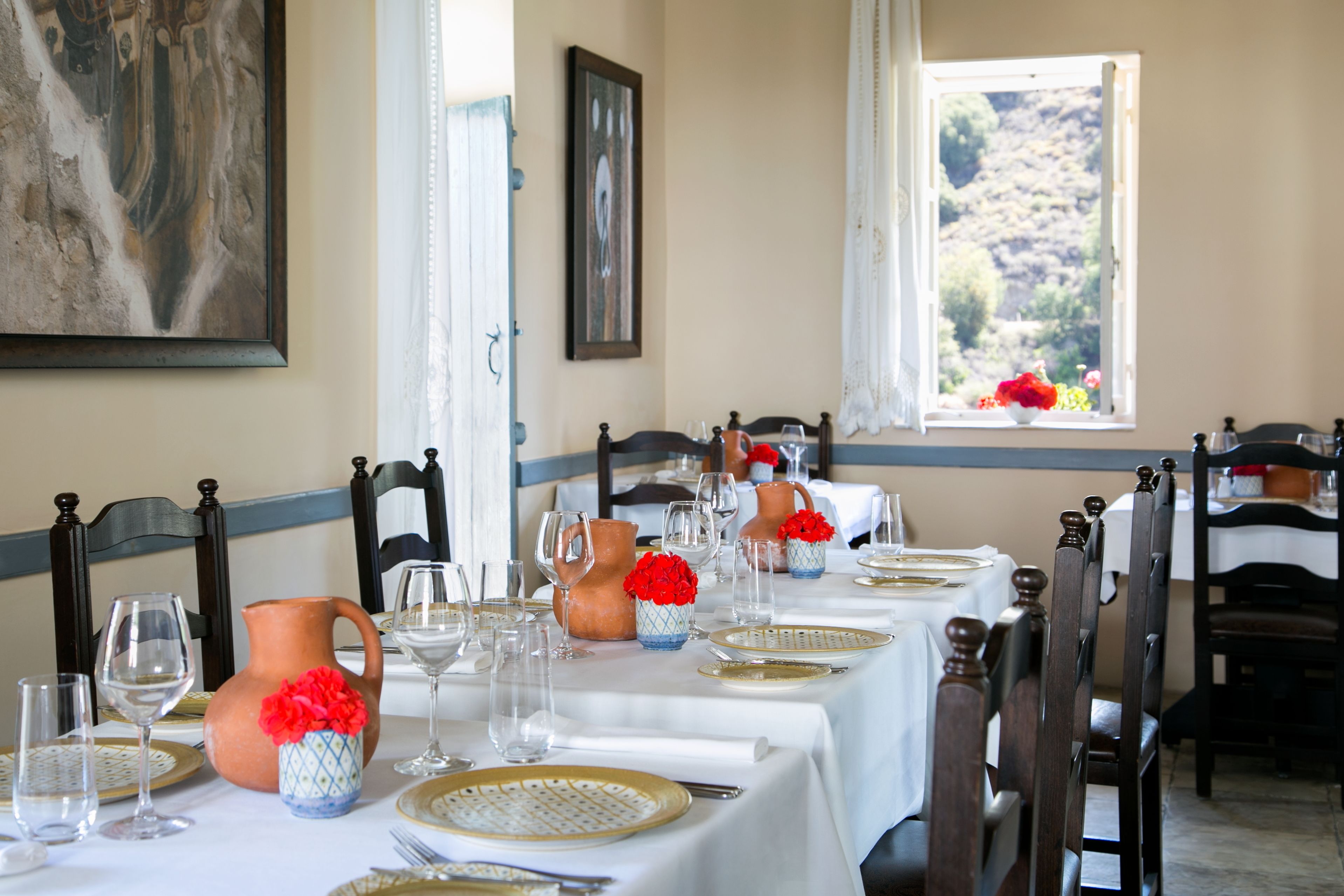 Une table à manger est dressée avec des nappes blanches, de l’argenterie dorée et blanche, et des verres à vin transparents. On y trouve des vases décoratifs avec des fleurs rouges et une cruche en terre cuite. La pièce est meublée de chaises en bois et possède une fenêtre donnant sur une colline.