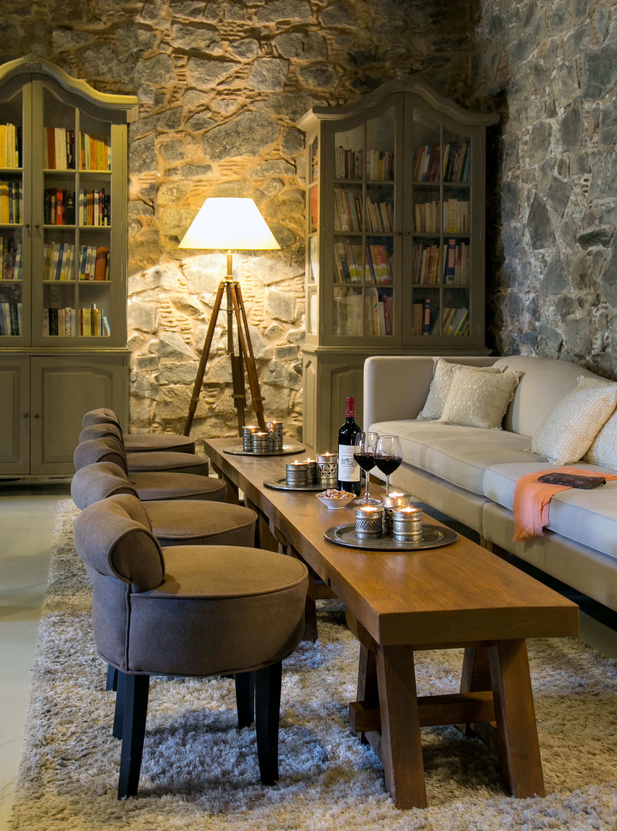 Uma sala de estar aconchegante com parede de pedra, mesa de centro de madeira, sofá e duas poltronas. Estantes repletas de livros e um abajur.