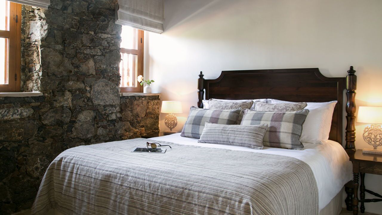 Un dormitorio con pared de piedra, cabecera de madera, cama con ropa de cama escocesa, dos mesitas de noche con lámparas y una ventana con vistas.