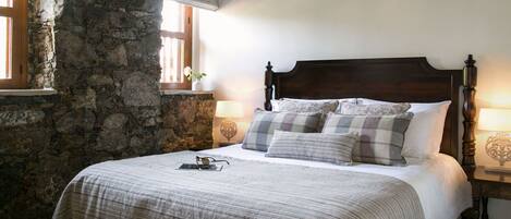 Un dormitorio con pared de piedra, cabecera de madera, cama con ropa de cama escocesa, dos mesitas de noche con lámparas y una ventana con vistas.