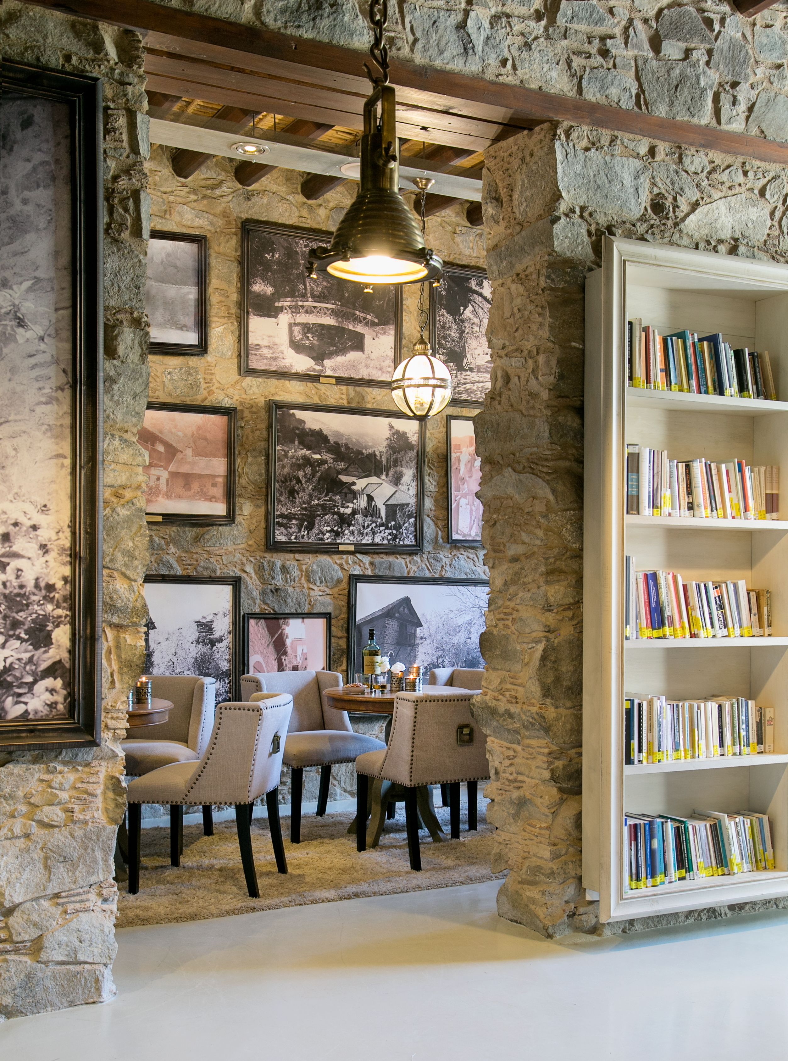 A cozy room with stone walls, a wooden ceiling, and a bookshelf filled with books. There are framed pictures on the walls and a table with chairs.