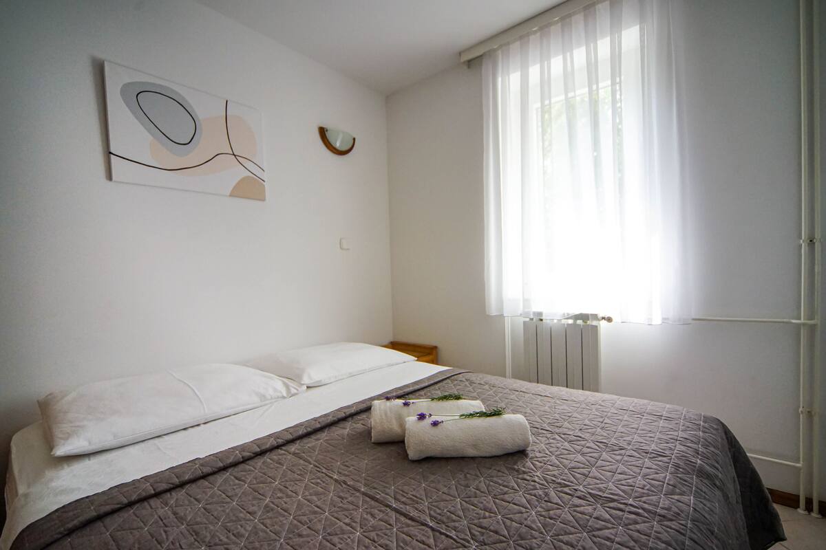 Ein ordentlich bezogenes Bett mit einer gesteppten Bettdecke, zwei Kissen und zwei Handtüchern mit Blumenmuster an einer weißen Wand mit abstrakter Kunst.