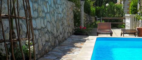 A swimming pool with stone walls and a wooden deck, surrounded by greenery and a stone wall.