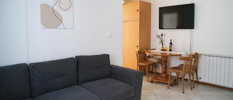 Una sala de estar con un sofá gris, una mesa de comedor con sillas, un televisor de pantalla plana montado en la pared y un radiador.