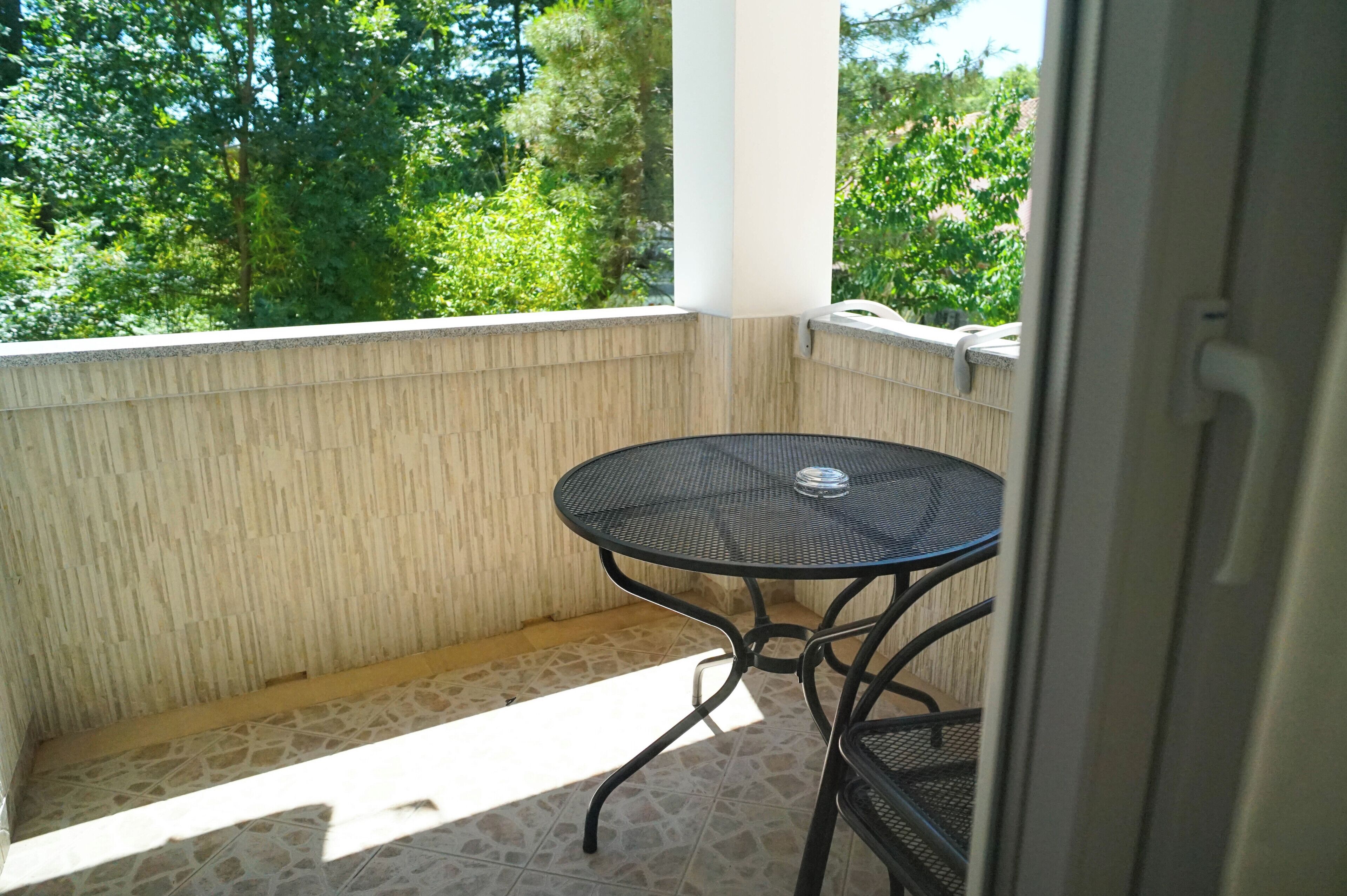 A balcony with a small round table and chairs, a tiled floor, and a view of trees.