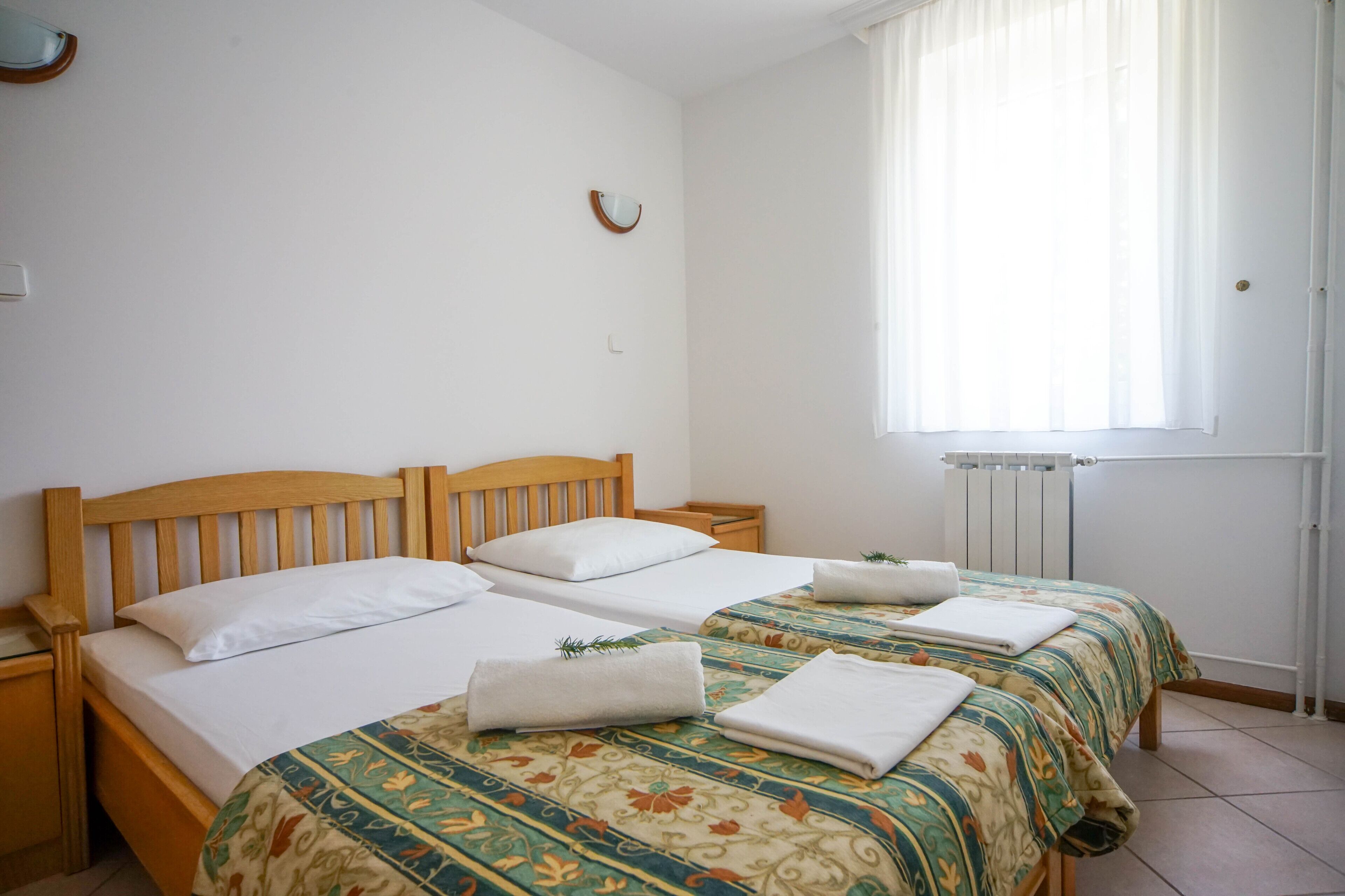 A double bed with a wooden headboard, two pillows, and a floral-patterned bedspread. There is a small bedside table with a lamp, and a window with sheer curtains.