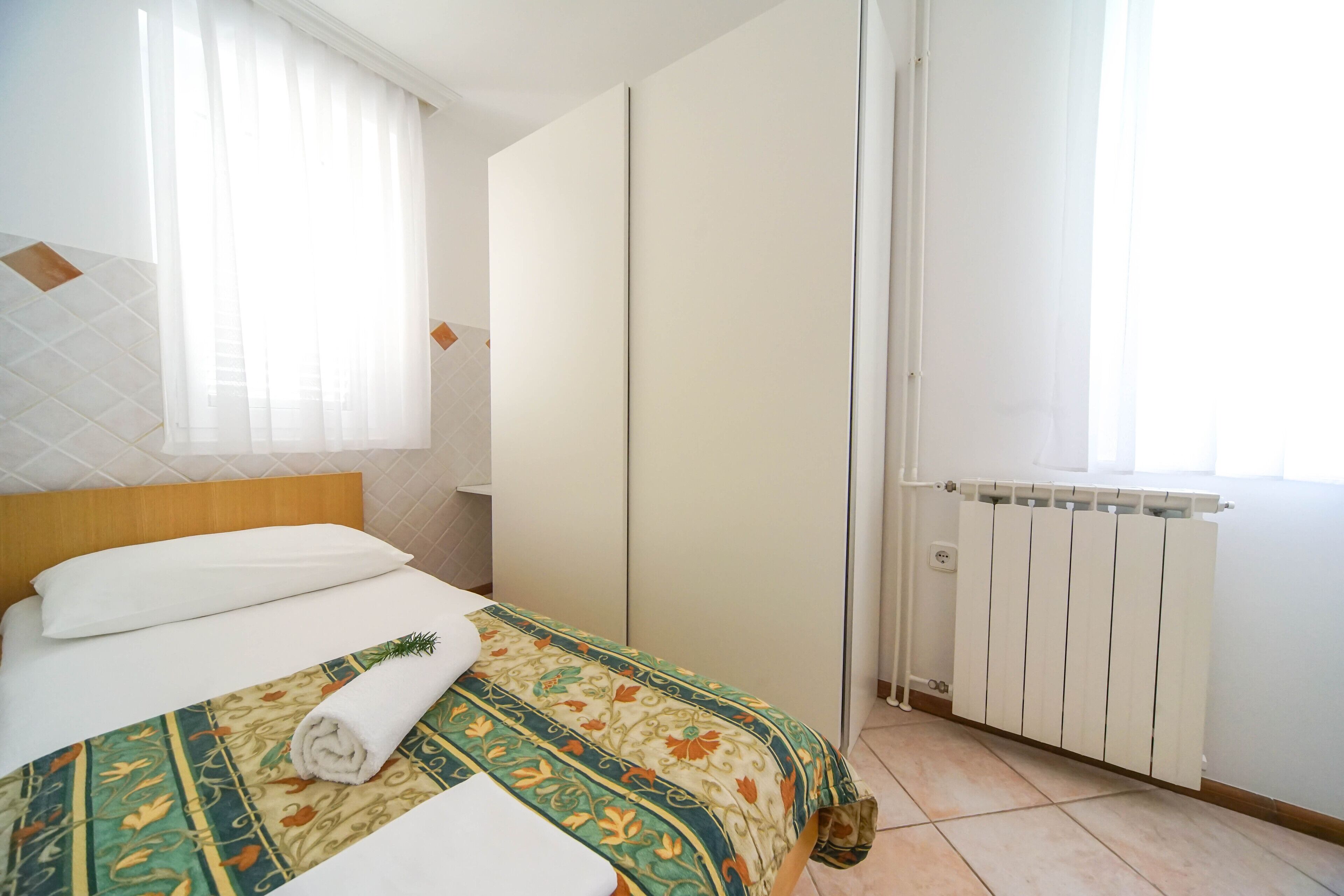 A hotel room with a neatly made bed, a radiator, and a window with curtains.
